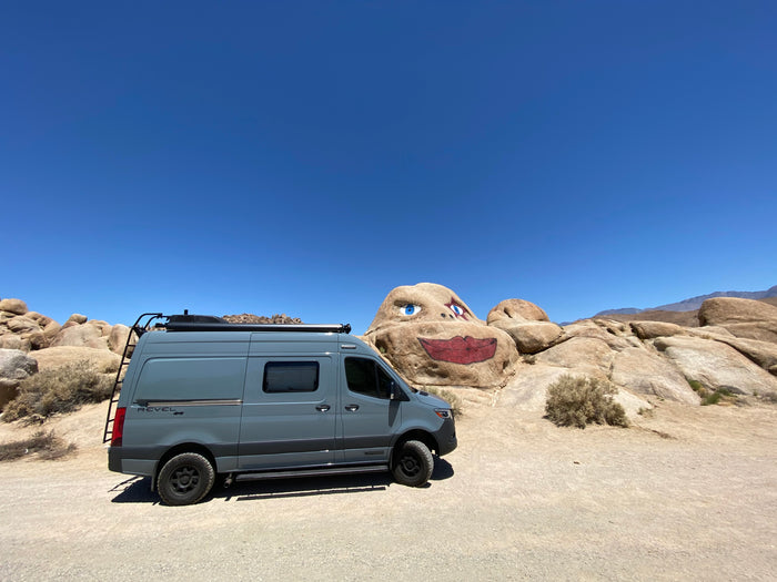 This grey Winnebago Revel 4x4 rv rental is parked on dirt, showcasing a unique painted rock face in the desert.