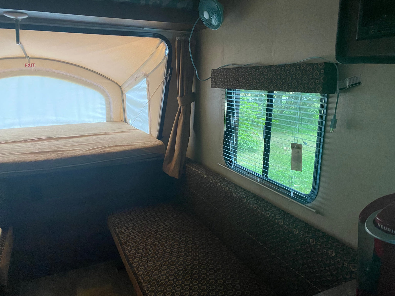 Interior of an RV rental showing a canvas pop-out bed, patterned dinette seating, and a window with blinds.