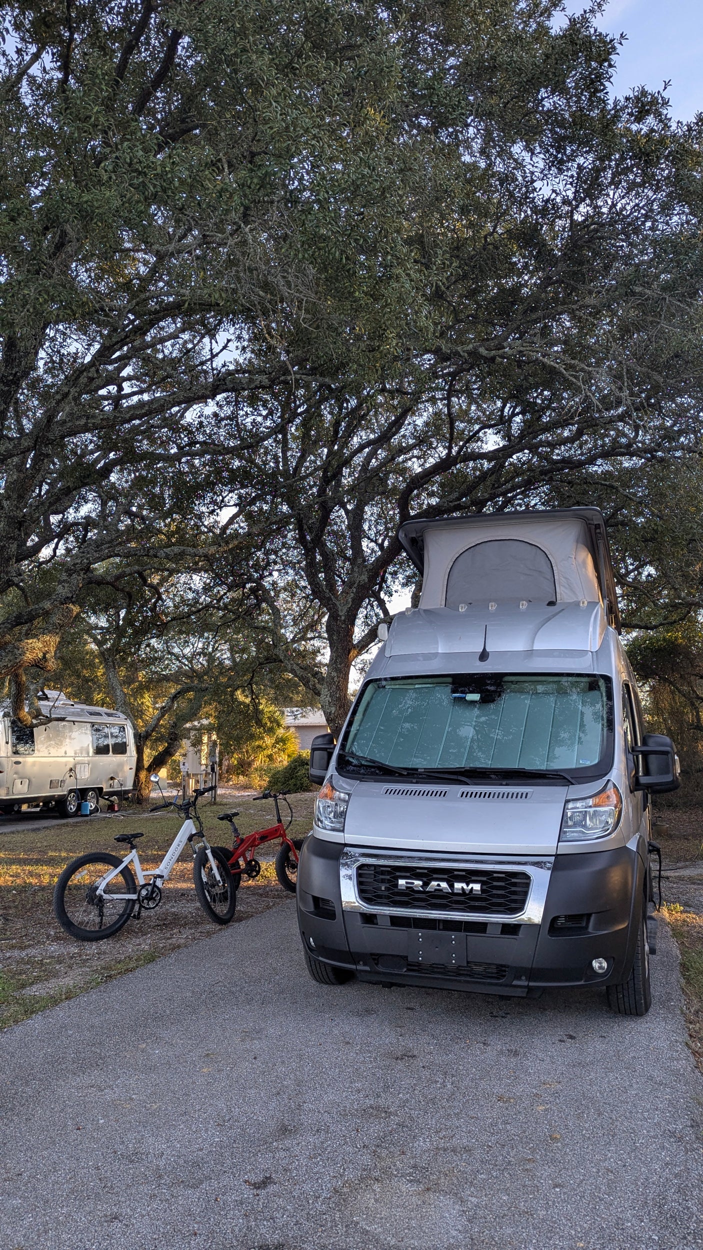 This RV rental camper van with a pop-top roof and two electric bikes is ready for adventure at a shady campsite.