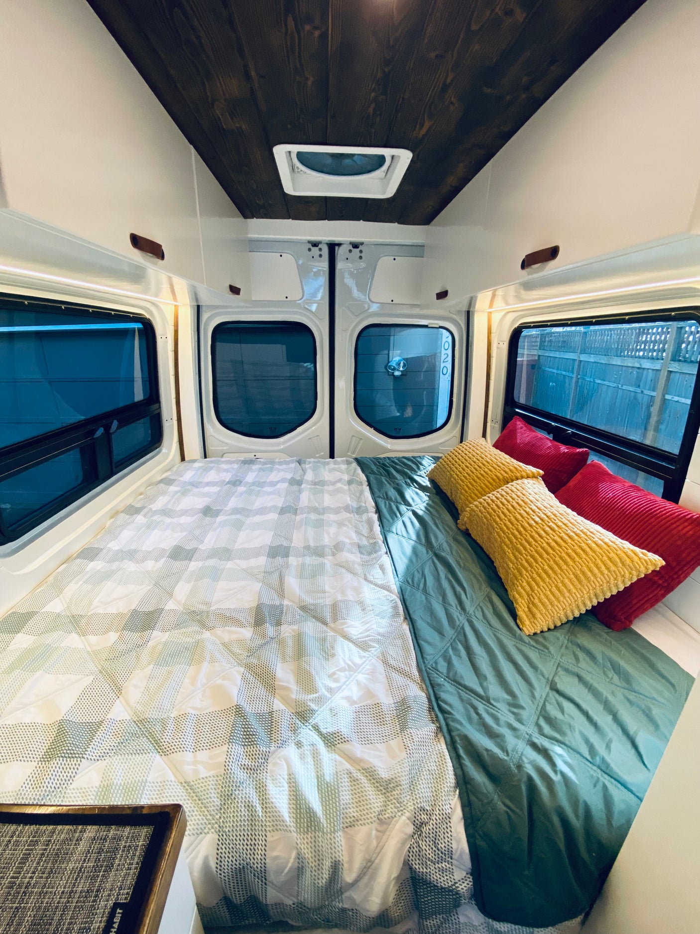 Inside a cozy RV rental: a comfortable bed with plaid/teal bedding, yellow/red pillows, wooden ceiling, and large windows.