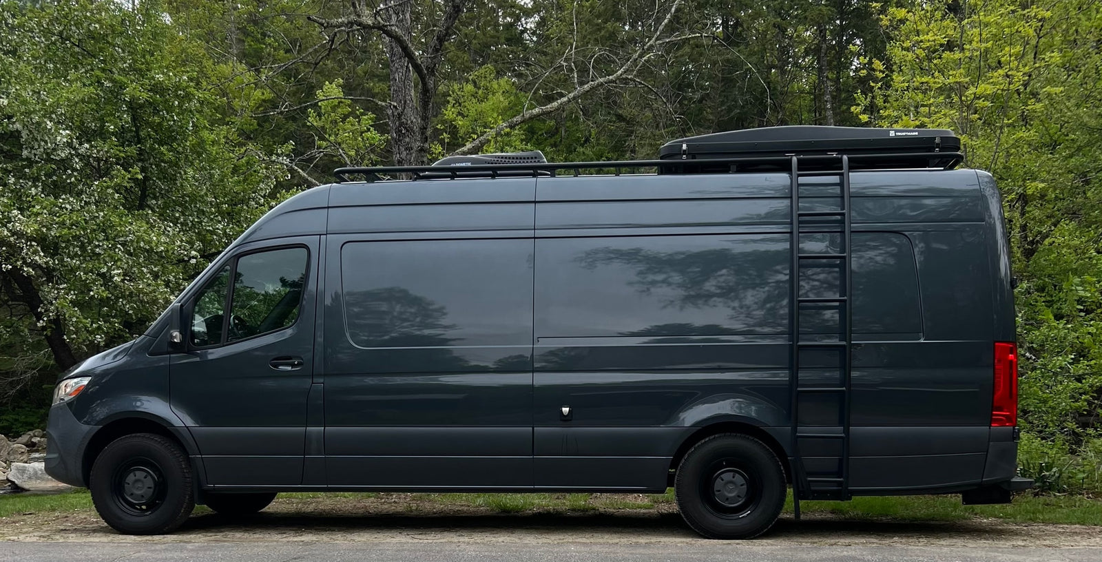 Explore with this dark gray Mercedes Sprinter RV rental. A custom camper van with roof rack & ladder, parked in nature.