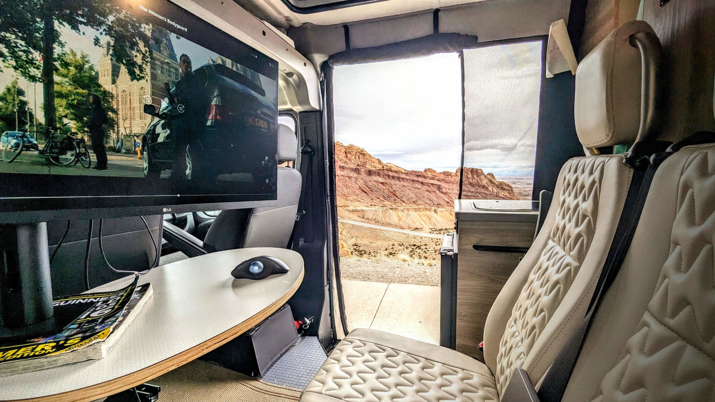 Inside an RV rental: a movie plays on screen, a desk with mouse is ready, and plush seats face a stunning desert view.