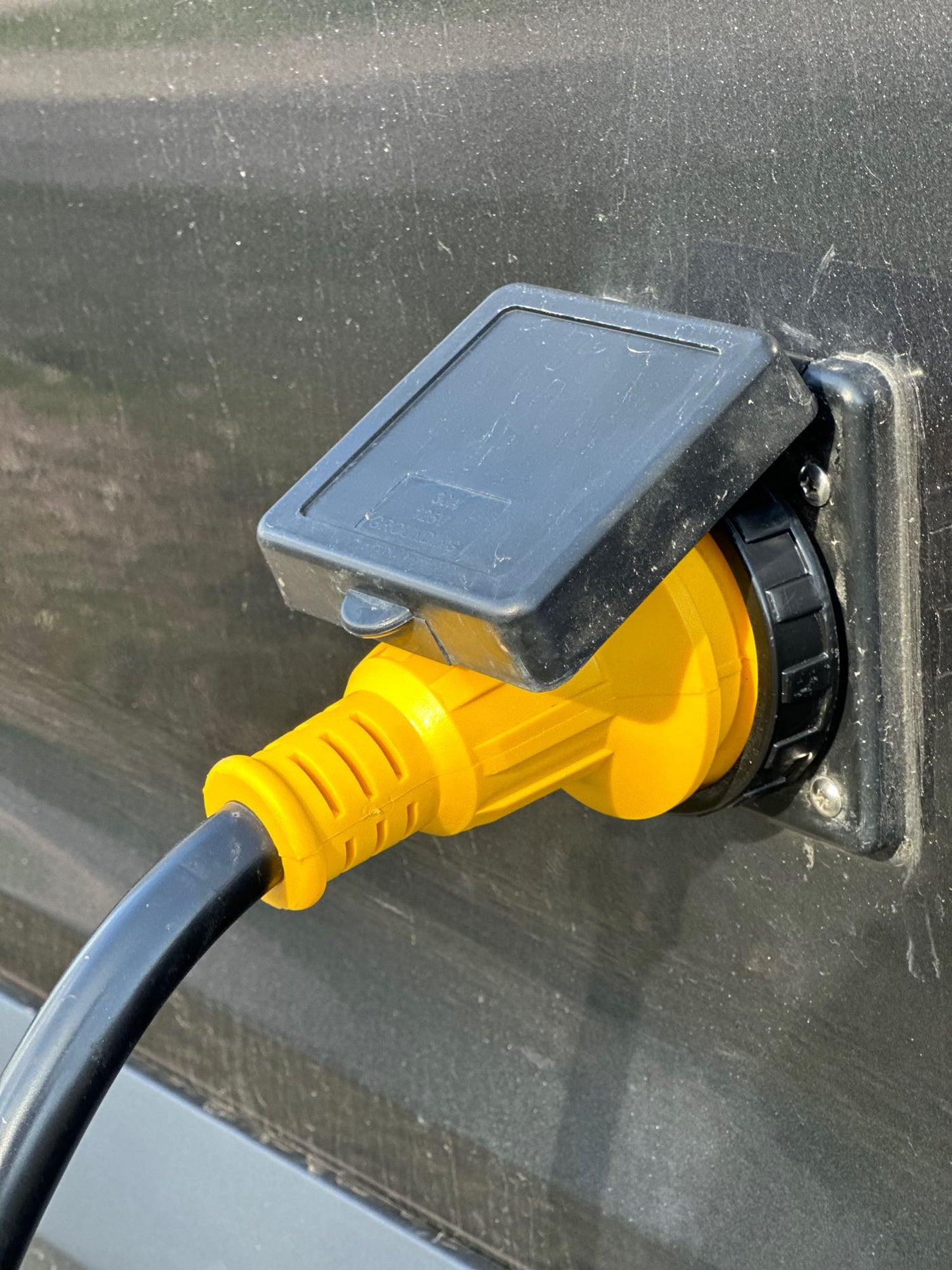 Bright yellow RV power cord plug inserted into a dusty black inlet on an RV rental, connecting to shore power.