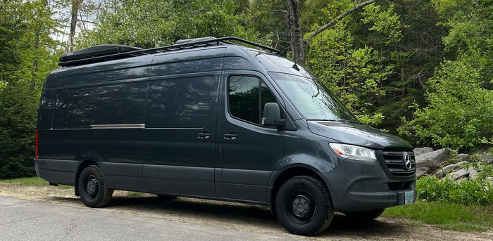 Dark gray Mercedes Sprinter camper van with roof rack & cargo box, ready for rv rental adventures, parked by a forest.