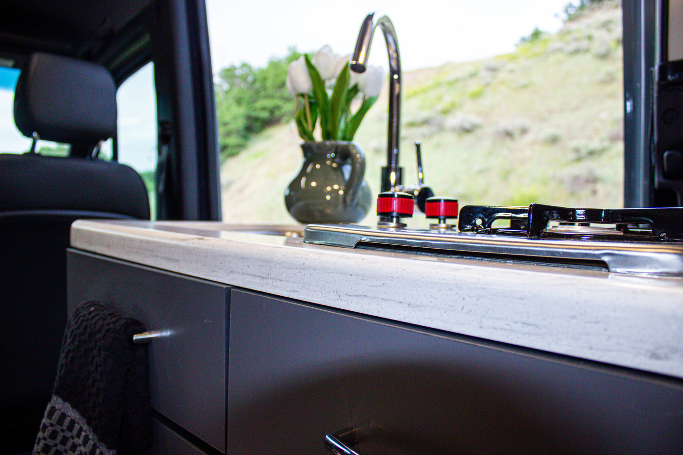 Modern rv rental kitchen with a sleek counter, sink, stovetop, and a vase of tulips, overlooking green hills.