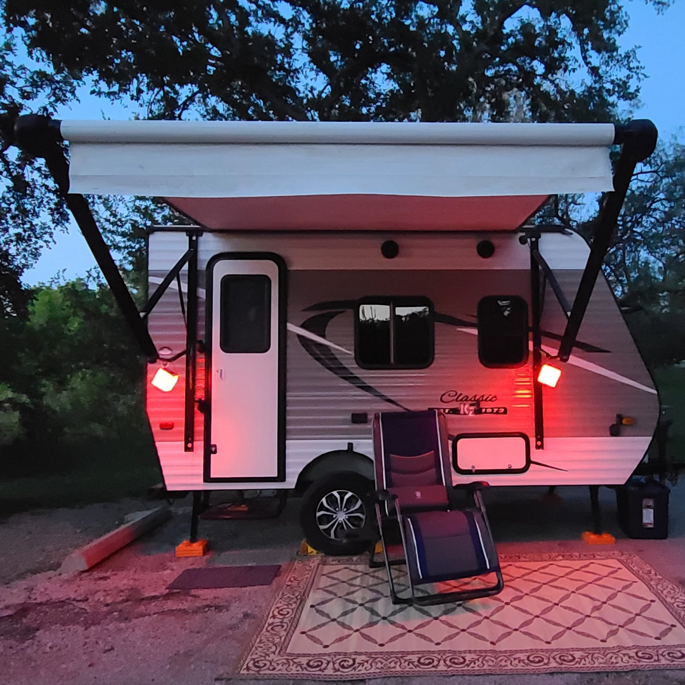 An rv rental camper at dusk/night, under an extended awning with red lights, a chair, and a patterned rug.