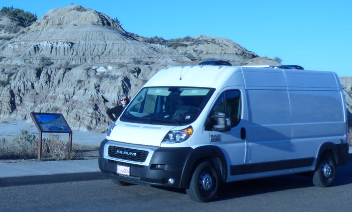 is parked near a calm blue body of water. A person stands on the left.|A white rv rental campervan parked near Devils Tower