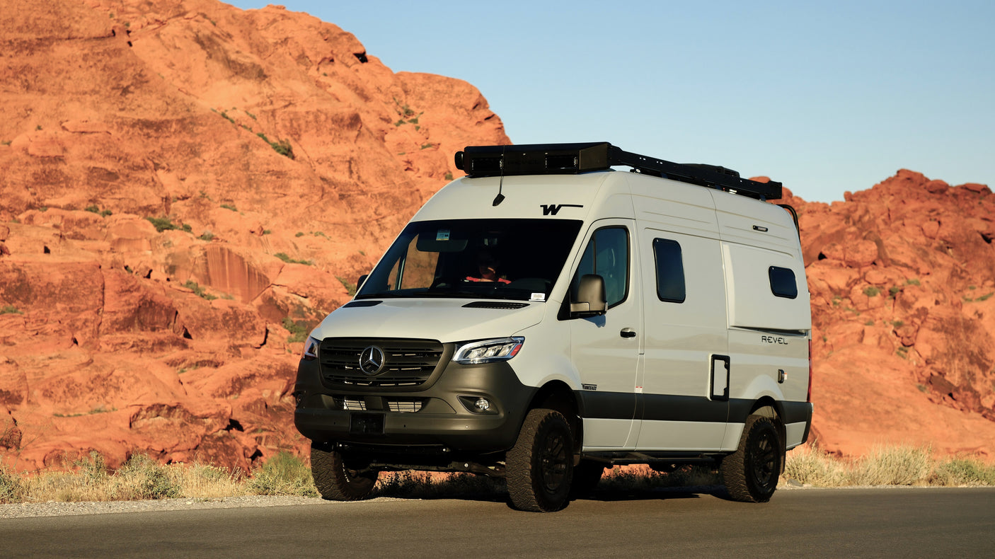 Winnebago Revel Sprinter rv rental, a light grey camper van, parked on a road near towering red rock desert mountains.
