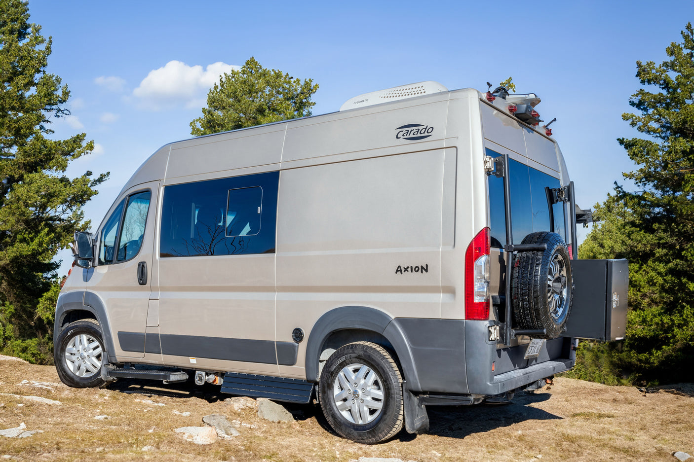 A tan Carado Axion camper van, equipped with a spare tire and roof AC, perfect as an RV rental for outdoor adventures.