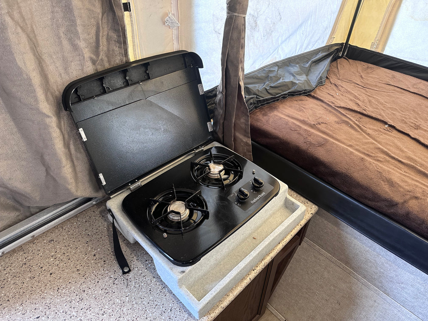 Interior of an RV rental pop-up camper, featuring a compact kitchen with a gas stove and nearby bed.