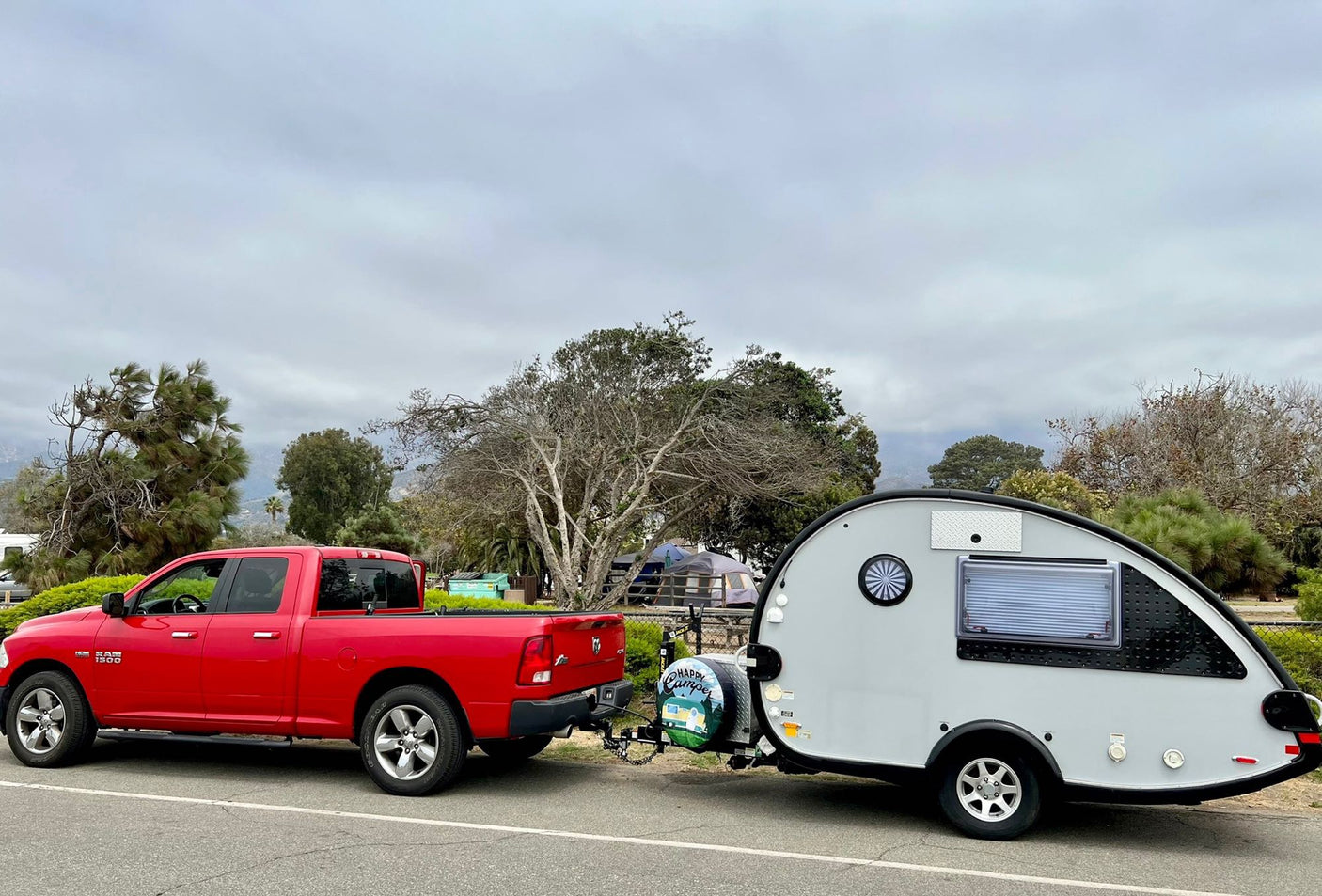 A red pickup truck tows a gray teardrop RV rental camper at a campsite under an overcast sky.