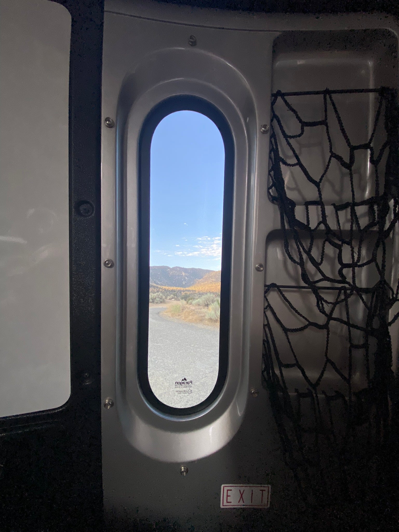 RV rental interior: oval window frames fall mountains, road. Storage nets & EXIT sign.