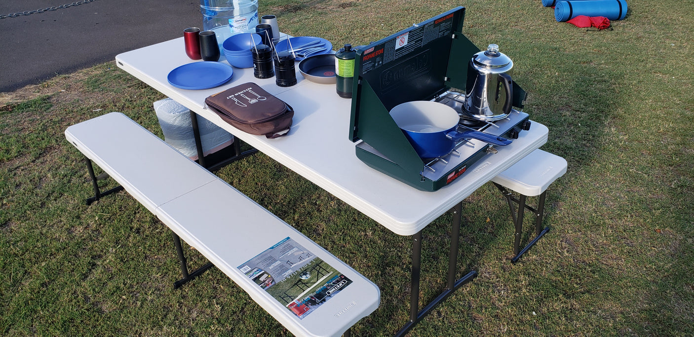 Portable camp kitchen setup on grass, perfect for an RV rental trip, featuring a table, benches, propane stove, and cooking essentials.