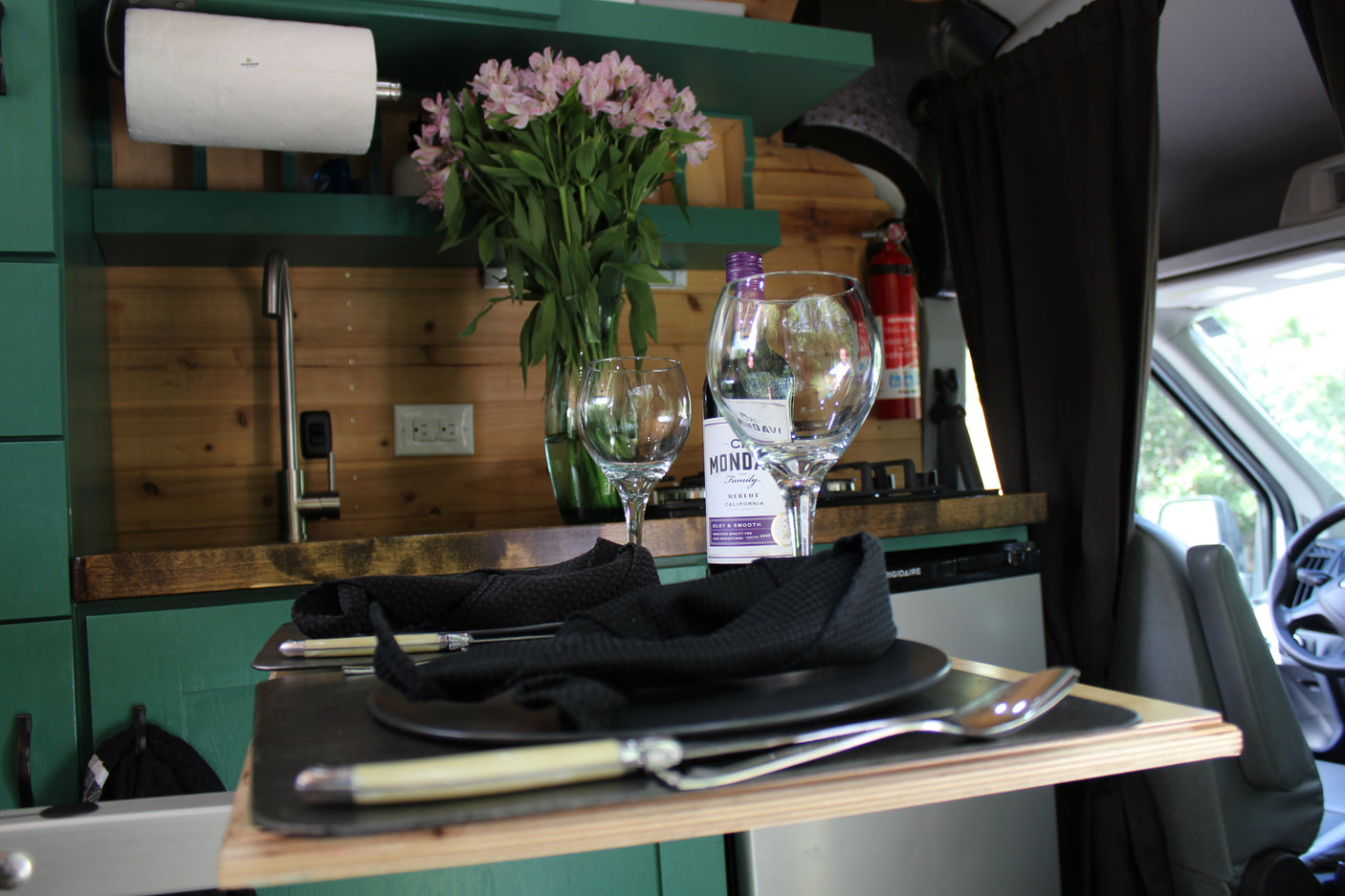 Cozy rv rental interior: wine, flowers, and dinnerware on a pull-out table. Kitchenette with sink and Frigidaire visible.