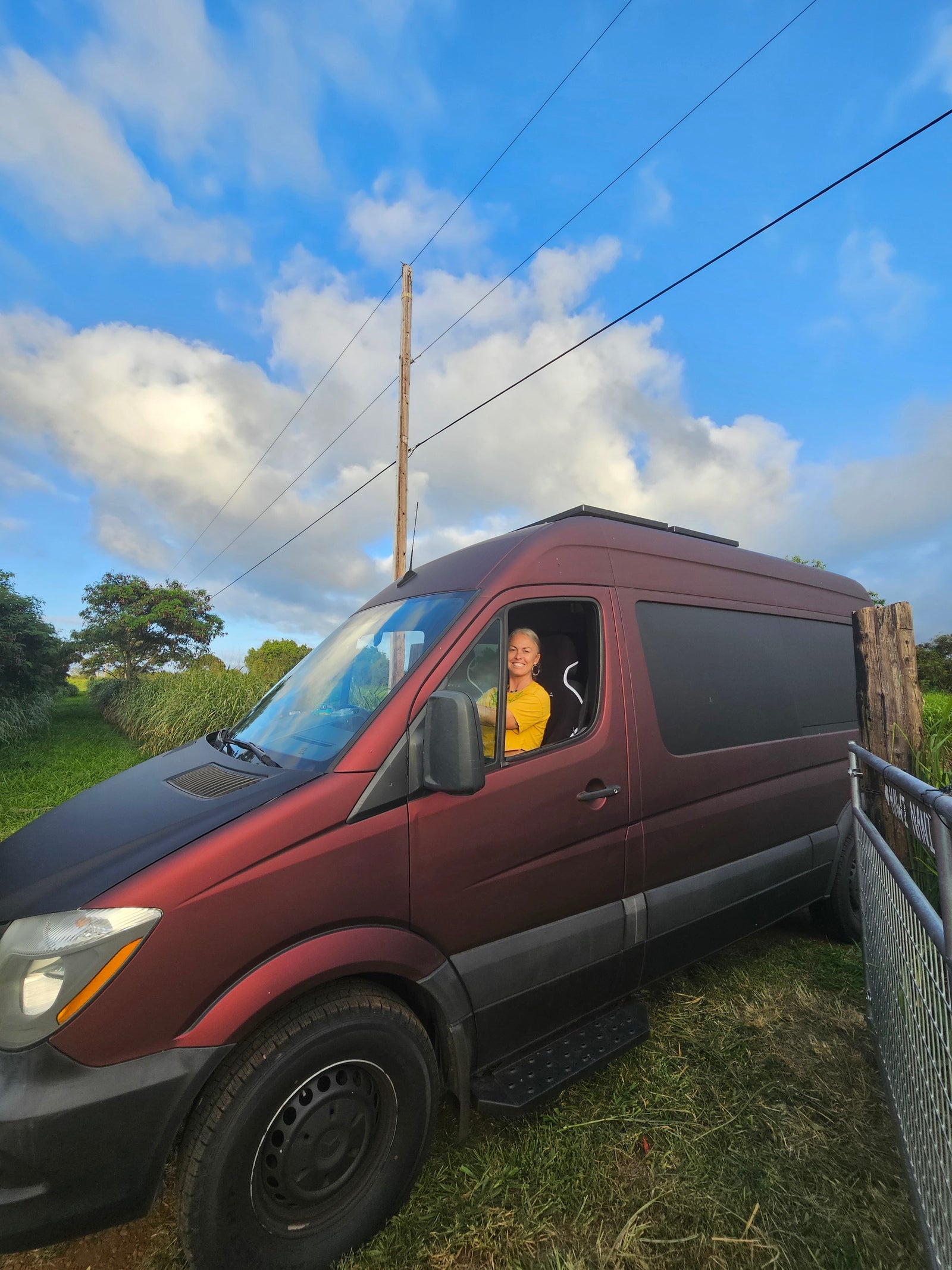 Smiling woman in a maroon rv rental van, parked in a grassy area under a bright blue, cloudy sky.