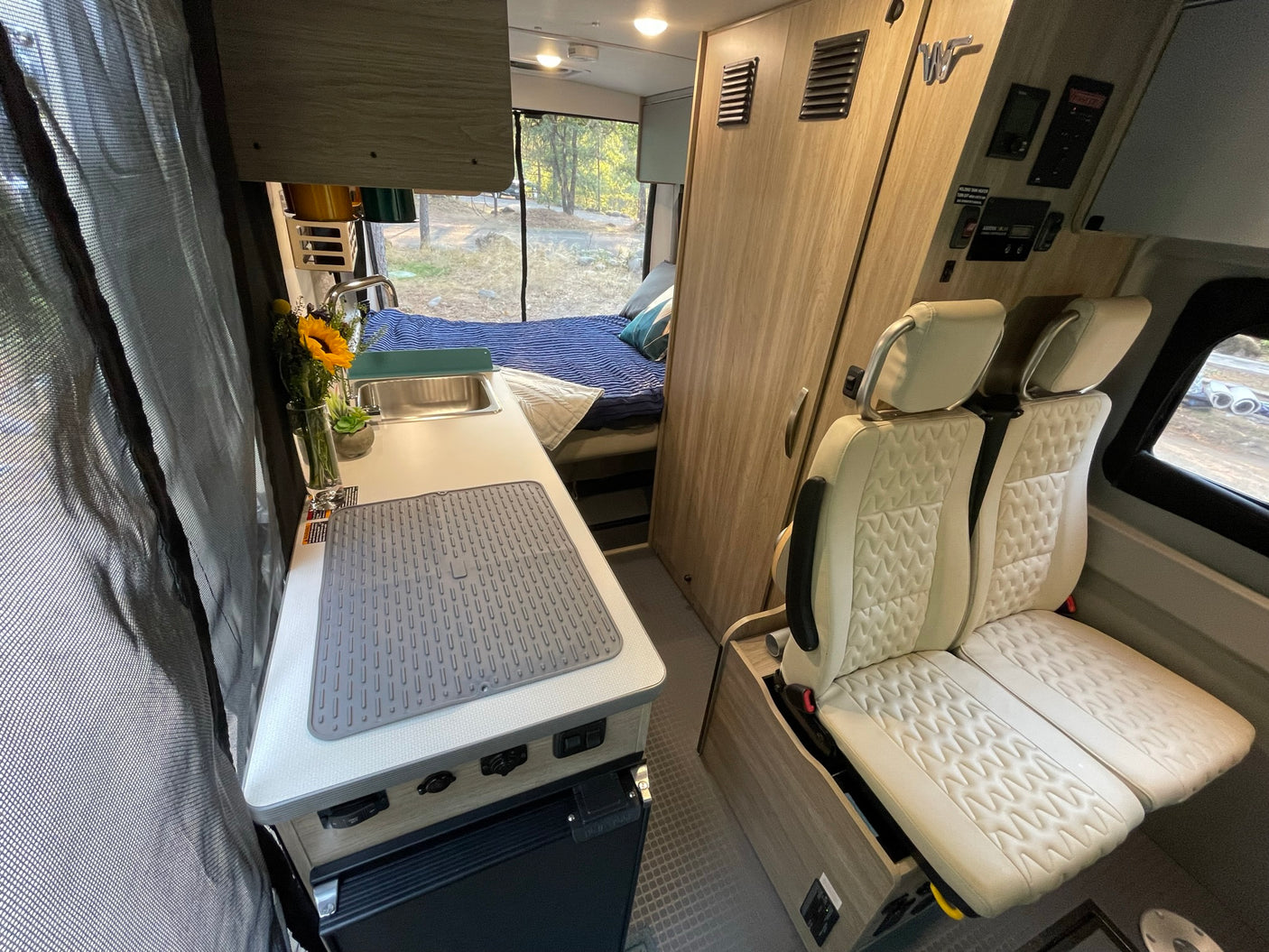 RV rental interior: A cozy camper van features a kitchen with sink, rear bed, and passenger seats, overlooking a campsite.