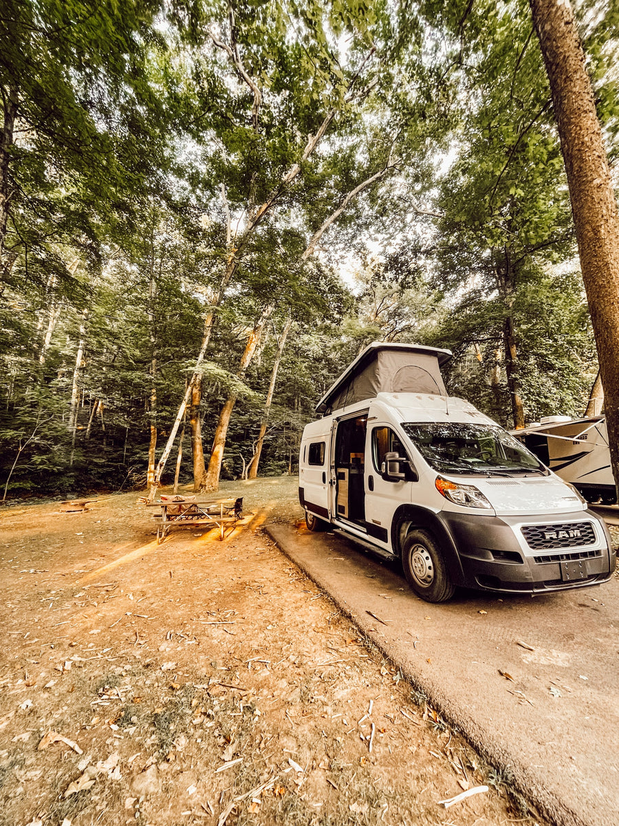 Experience nature with an RV rental! A white camper van is parked at a forested campsite with a picnic table and fire pit.|White camper van with pop-top & open door on green grass