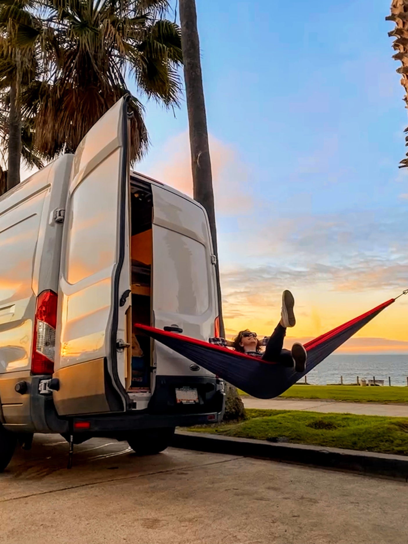 RV rental: Person relaxes in hammock attached to white camper van, enjoying ocean sunset views. Ideal van life adventure.