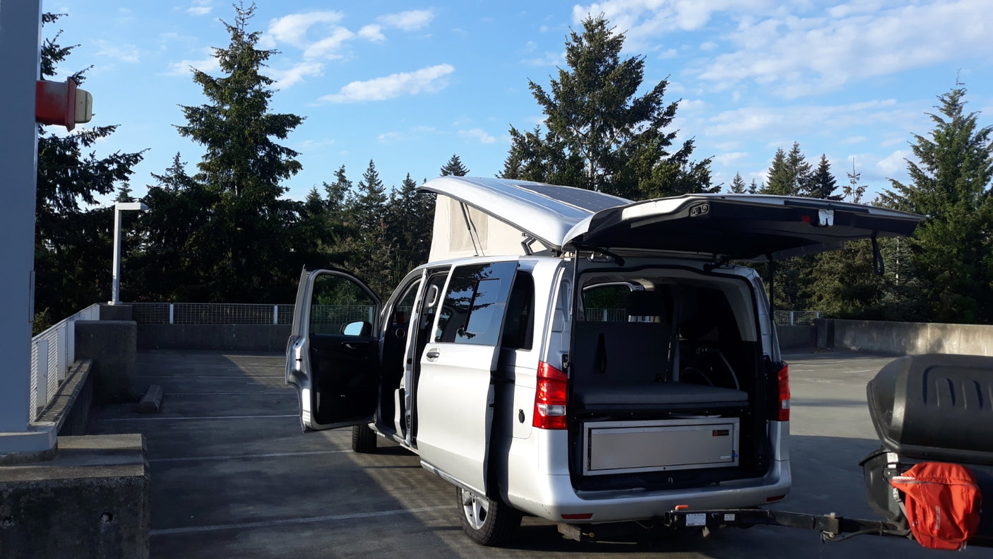 Silver pop-top RV rental camper van with open doors and a cargo box. Parked in a concrete lot amidst green trees.