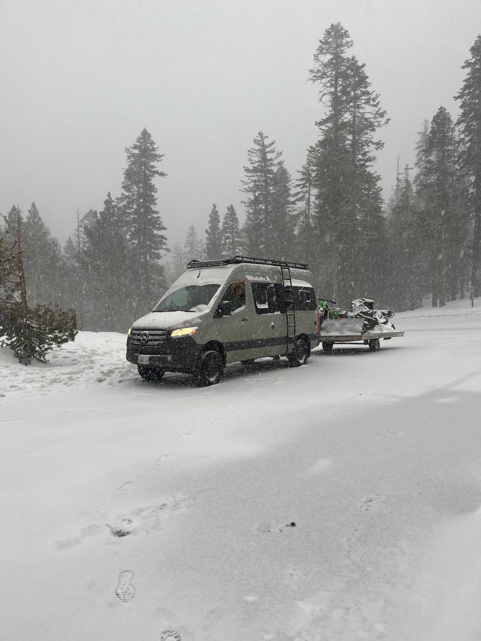 A gray Mercedes-Benz RV rental with a roof rack is towing snow-covered snowmobiles through a snowy forest.