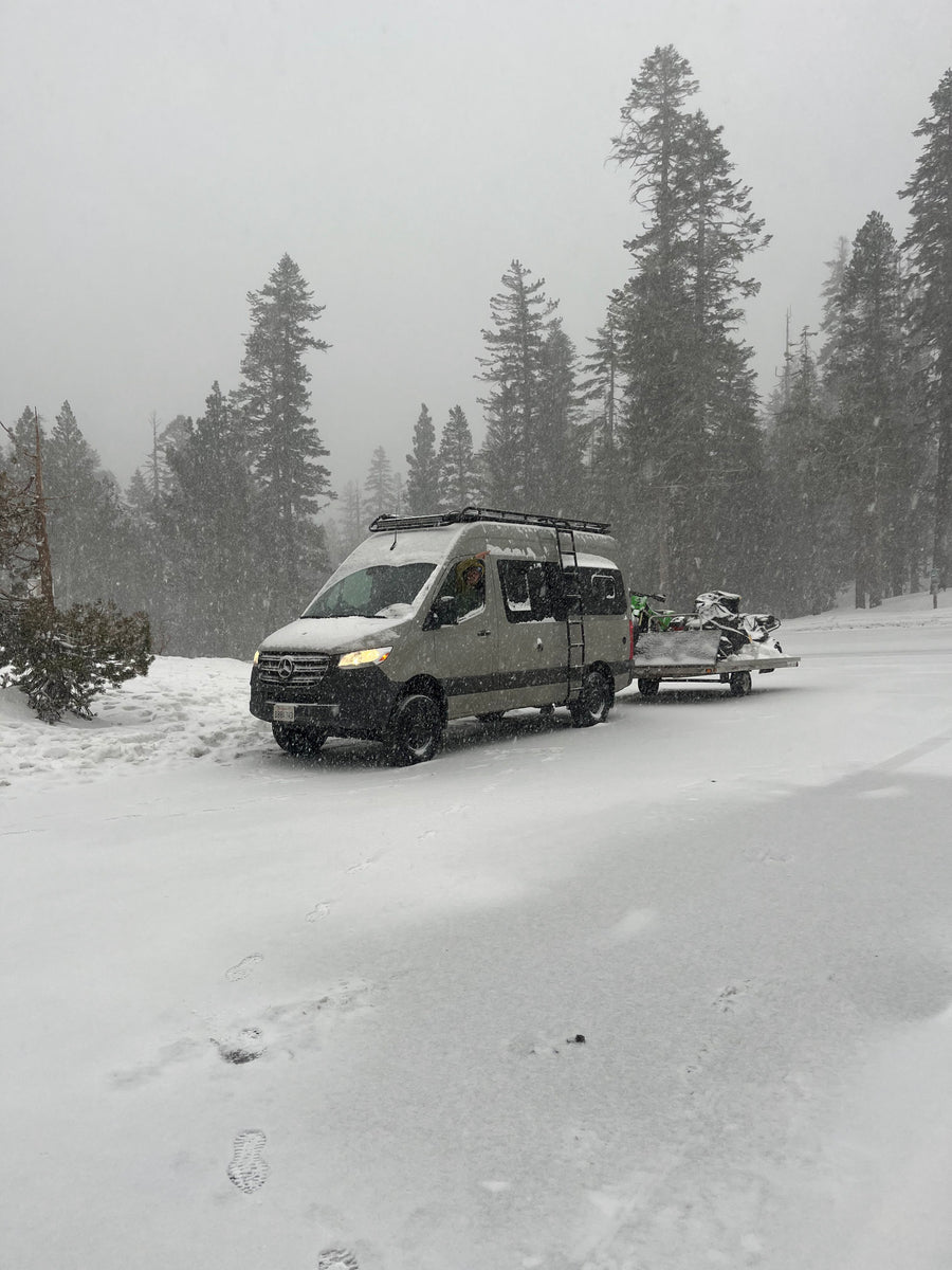 A gray Mercedes-Benz RV rental with a roof rack is towing snow-covered snowmobiles through a snowy forest.
