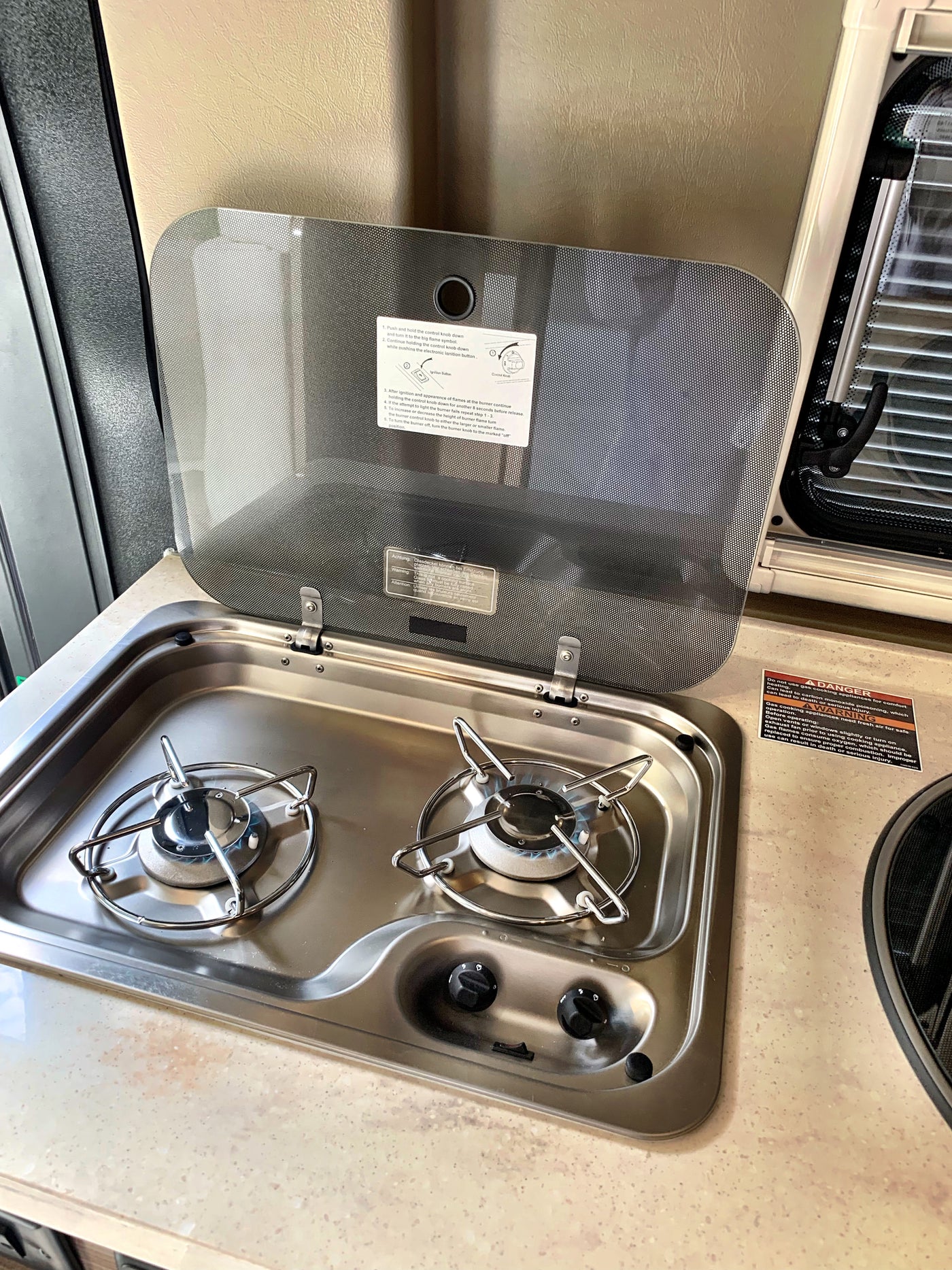 RV rental kitchen featuring a two-burner gas cooktop with an open safety lid, showing usage instructions and a lit burner. — photo 7