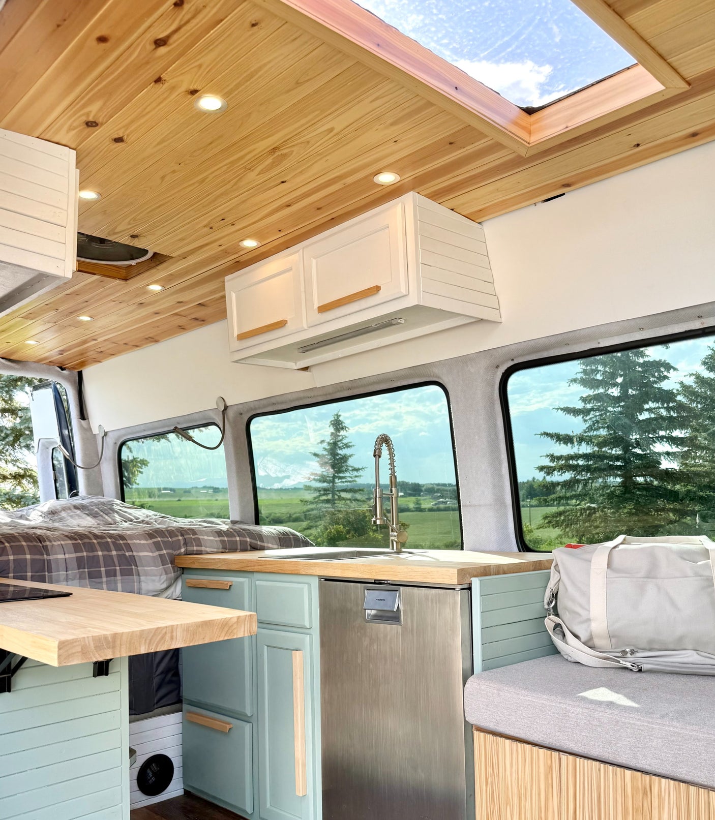 This rv rental's spacious camper van interior features a wood ceiling, skylight, light blue kitchen, and a bed with scenic views.