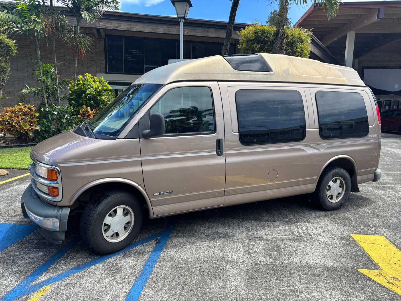 Tan Chevy 1500 Discovery high-top van, perfect for rv rental, parked by a building and tropical plants.