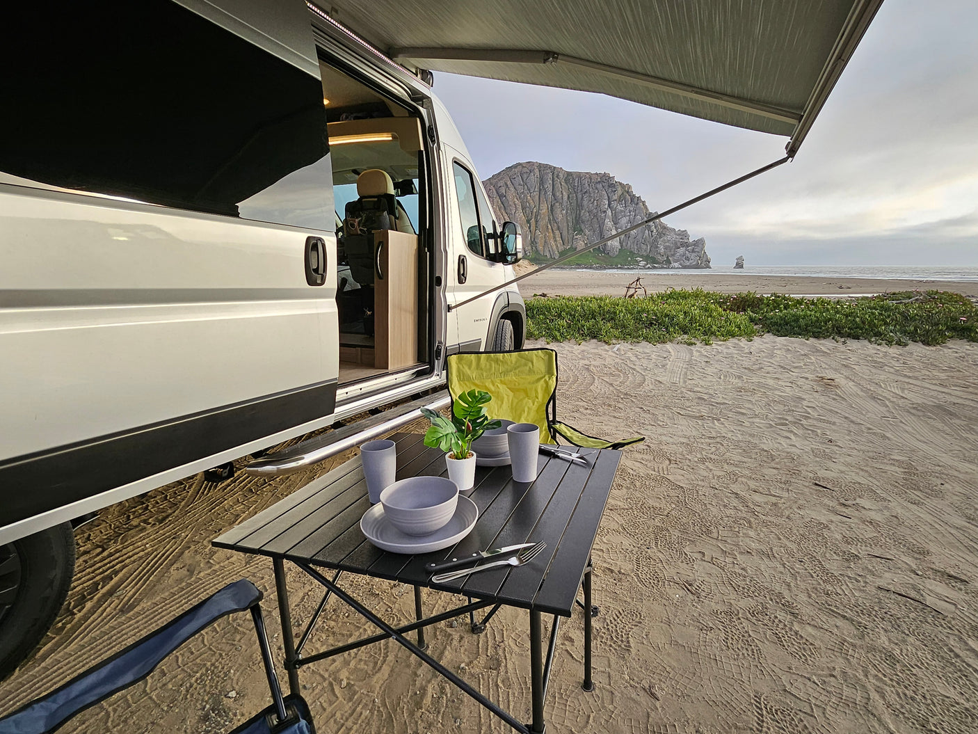 Beachside living with an **rv rental**: A camper van parked on sandy beach with a table set up, facing a mountain and ocean.