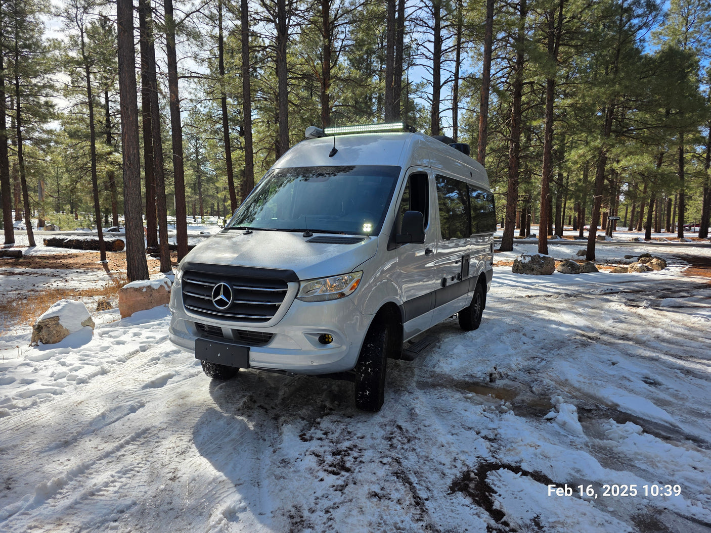 RV rental: A silver camper van parked in a snowy pine forest, surrounded by tall trees and white snow. Winter scene.