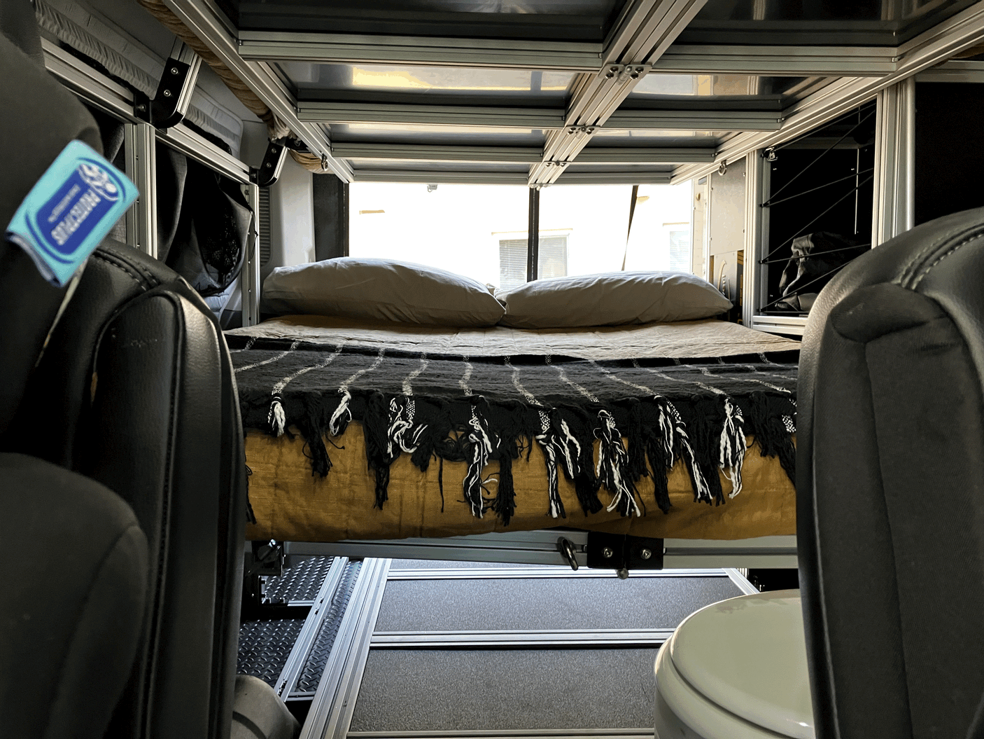 Cozy RV rental interior: A view of the elevated bed with pillows and a blanket, showing a modular frame, front seat, and toilet.