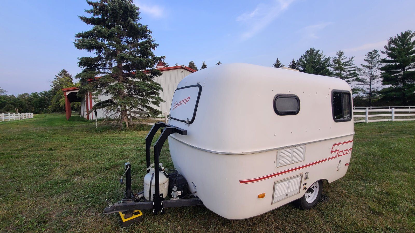 This compact white Scamp travel trailer (RV rental) is parked on grass, with a barn and pine trees in the background.