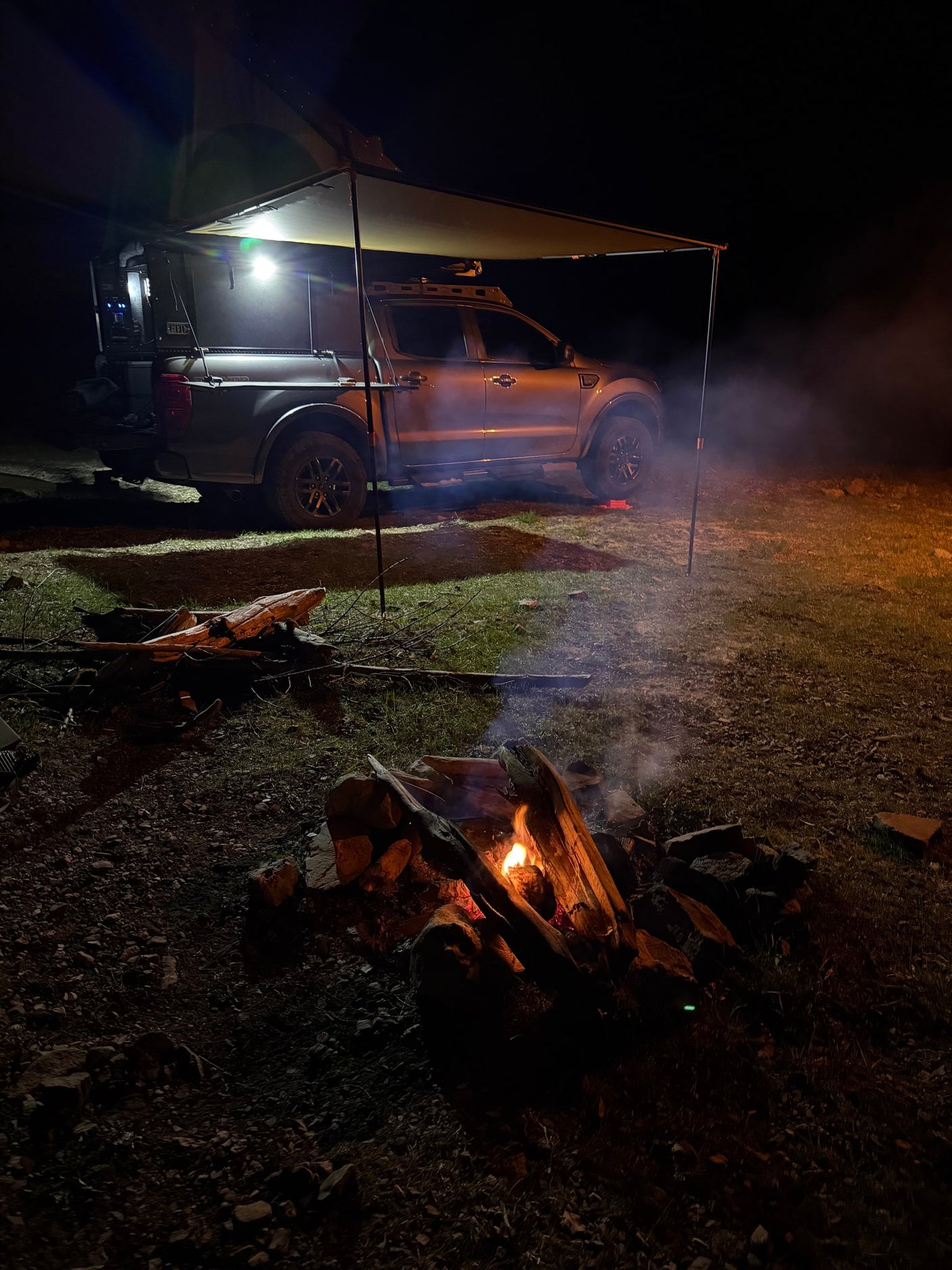 Night camping: A truck with an awning & bright lights next to a cozy campfire. Experience overland adventure with an RV rental!
