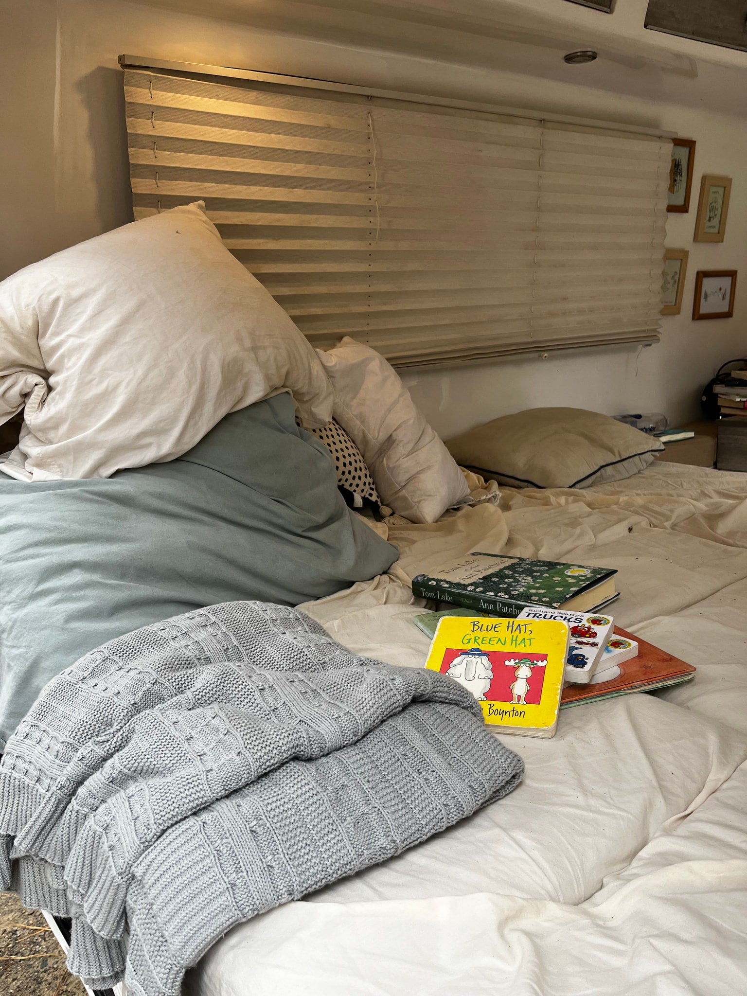 Cozy rv rental bed with white and light blue pillows, grey knitted blanket, a stack of books, and horizontal window blinds.