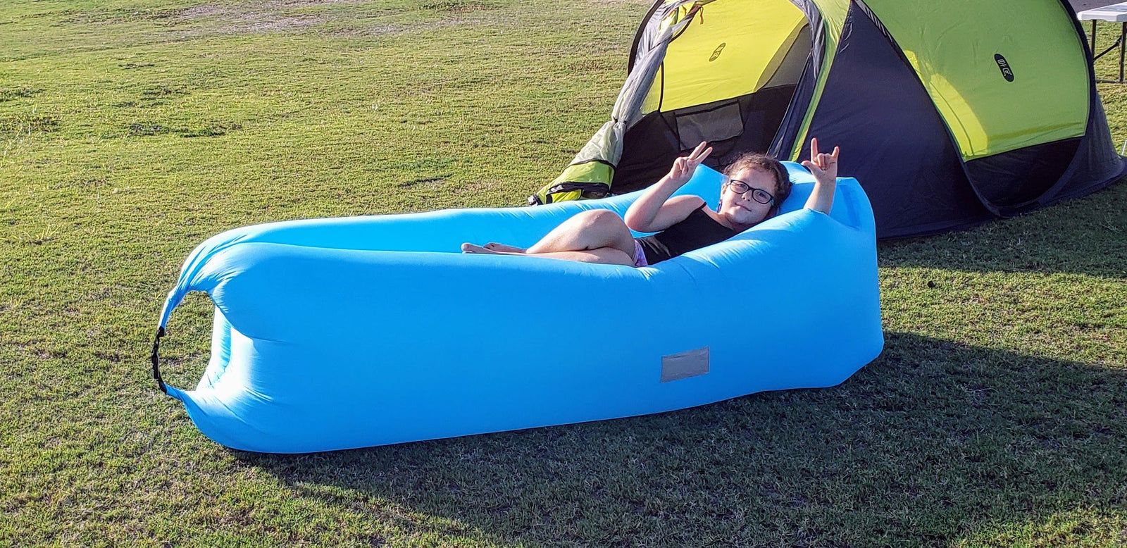 Young person enjoys a blue inflatable lounge chair by a green tent. Relaxing outdoors is ideal for your next RV rental adventure!