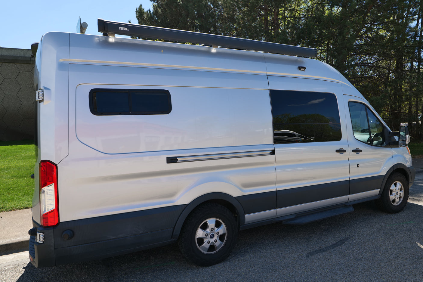 Silver Ford Transit RV rental van with a Thule awning, custom windows, and off-road tires parked outdoors.