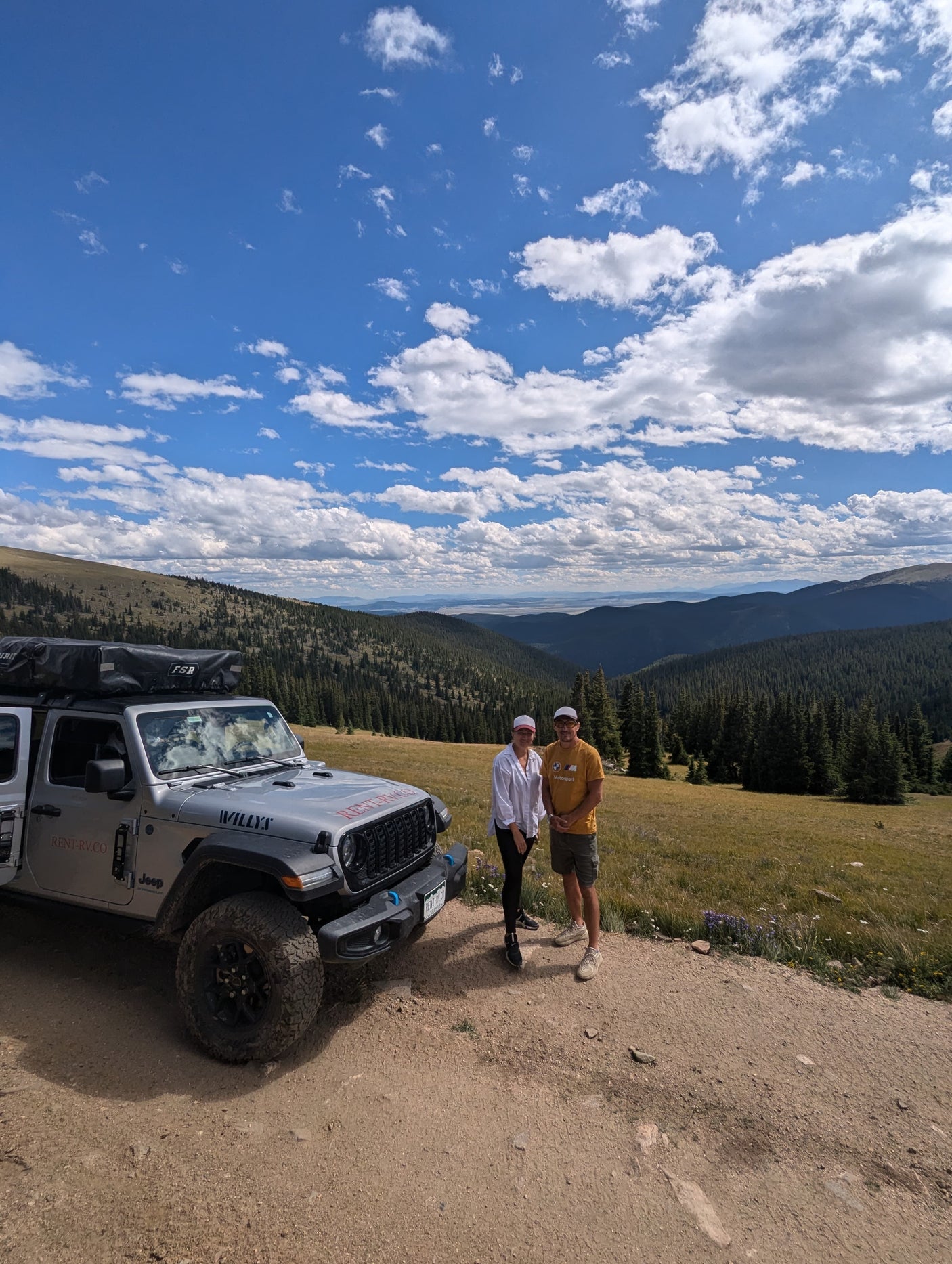 Enjoying the scenic views! A silver Jeep Wrangler **RV rental** with a rooftop tent on a mountain road, with a couple and vast landscapes.