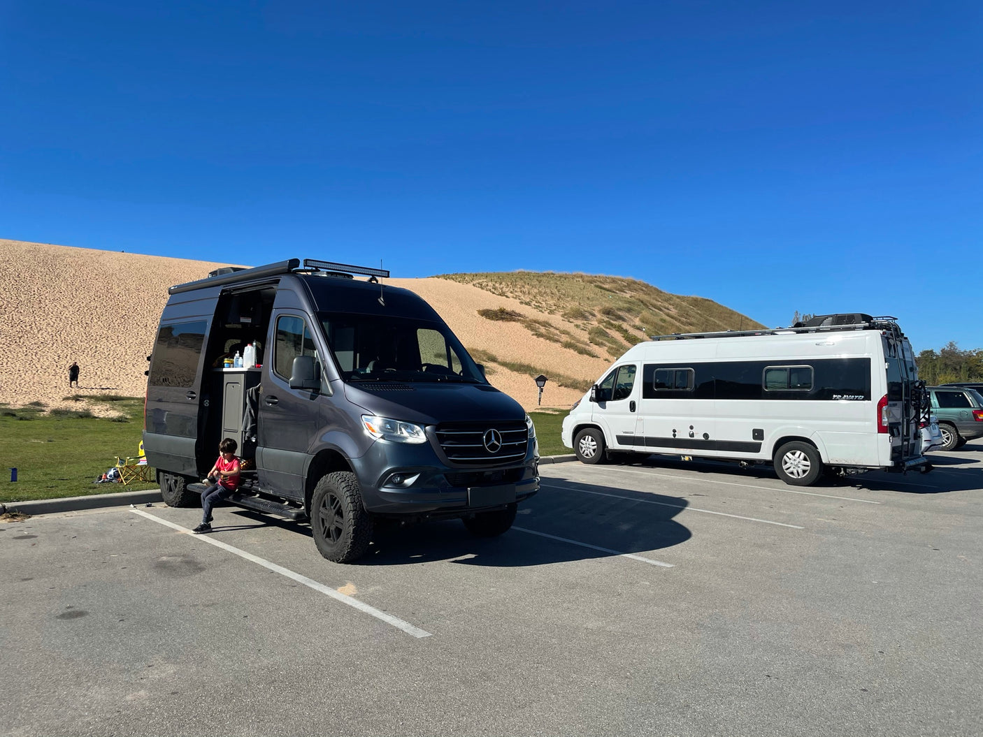 Experience van life! Two custom camper vans, resembling popular RV rental options, parked by sandy dunes. A child sits nearby.