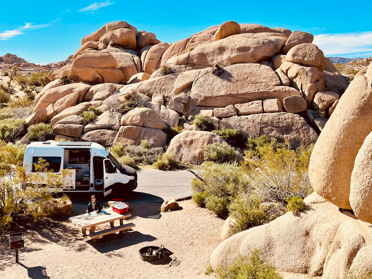 A scenic rv rental adventure at a desert campsite, featuring a camper van, person at a picnic table, and massive rock formations.