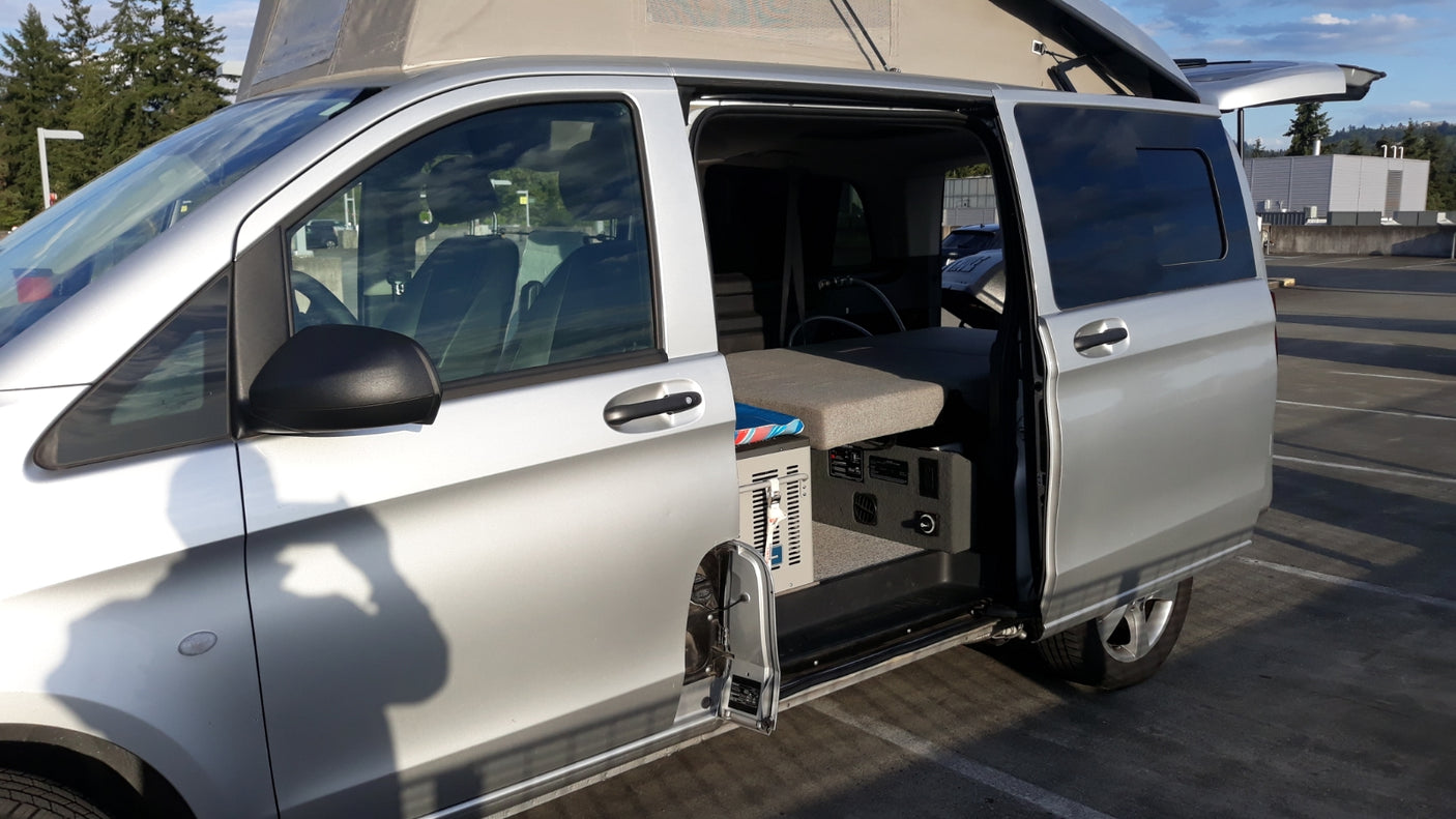 Silver camper van with pop-top roof and open side door revealing its compact interior setup, great for an RV rental.