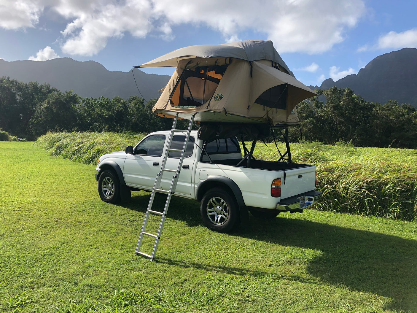 White pickup truck with a deployed tan rooftop tent & ladder on green grass, ready for your RV rental mountain adventure.