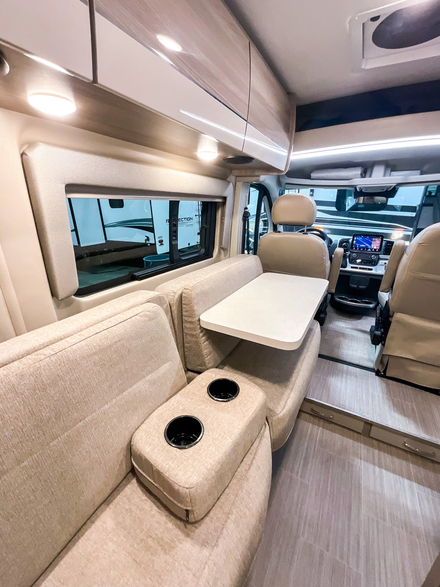 RV rental interior features a cream dinette, table, and cup holders. Bright overhead lighting and driver's area visible.
