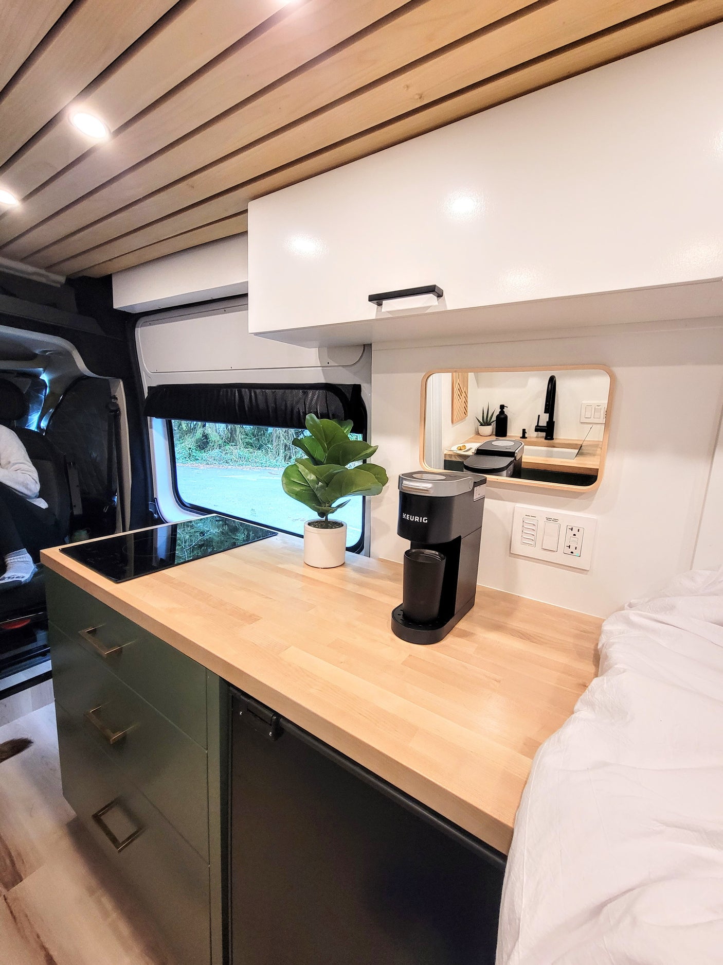 Bright RV rental interior featuring a compact kitchen with wood countertops, induction cooktop, and coffee maker, plus a bed.