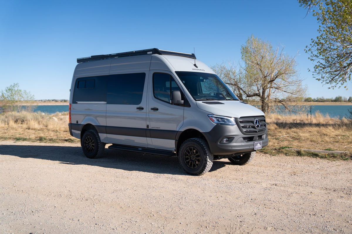 Light gray Mercedes Sprinter campervan rv rental with off-road tires, parked on a gravel path by a lake. — photo 1