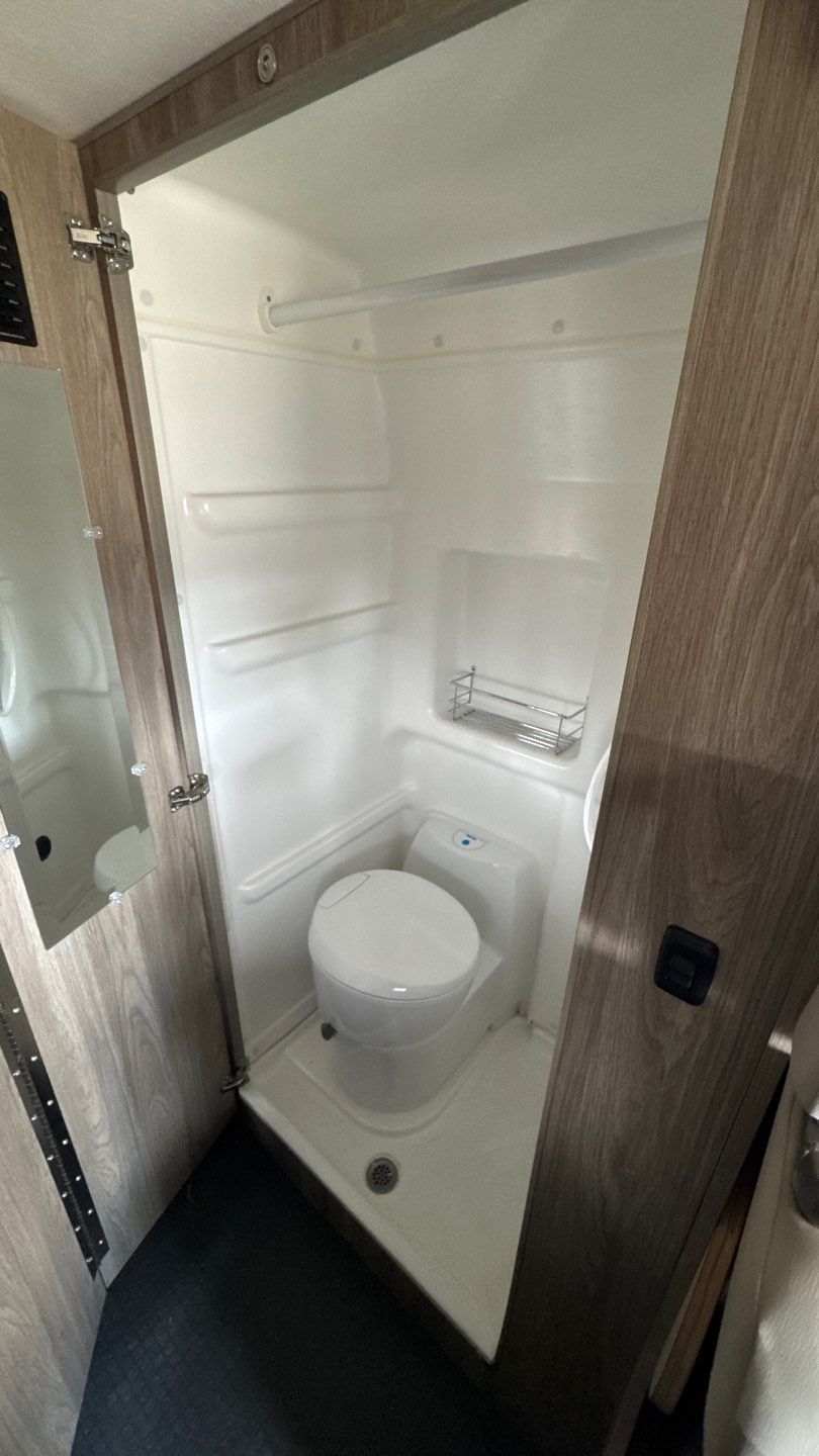 An RV rental's compact wet bath showing a white toilet, shower pan, and wall-mounted storage rack within the integrated shower space.