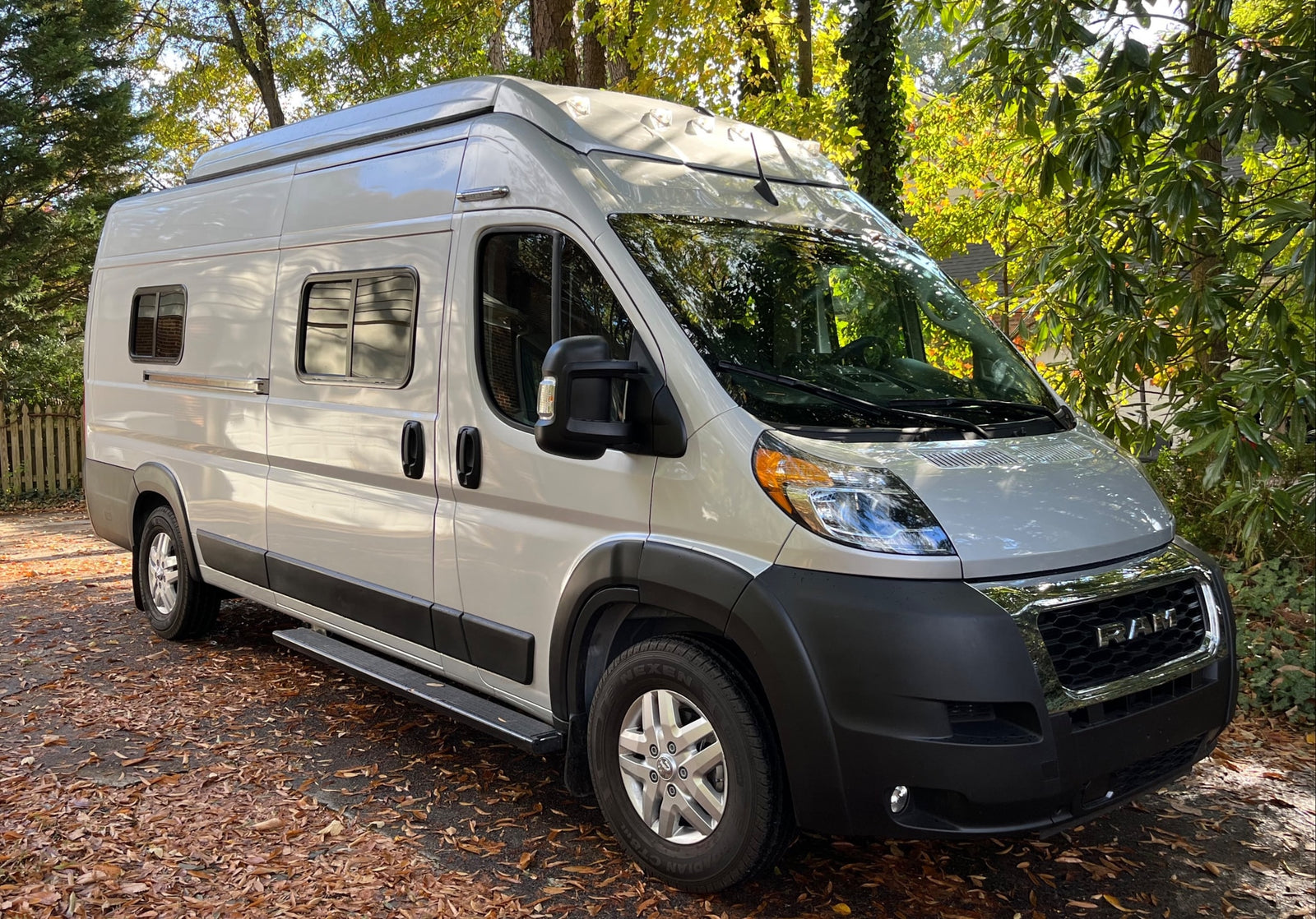 A light-colored Ram ProMaster camper van, an ideal RV rental, parked on a leafy autumn driveway with trees in the background.