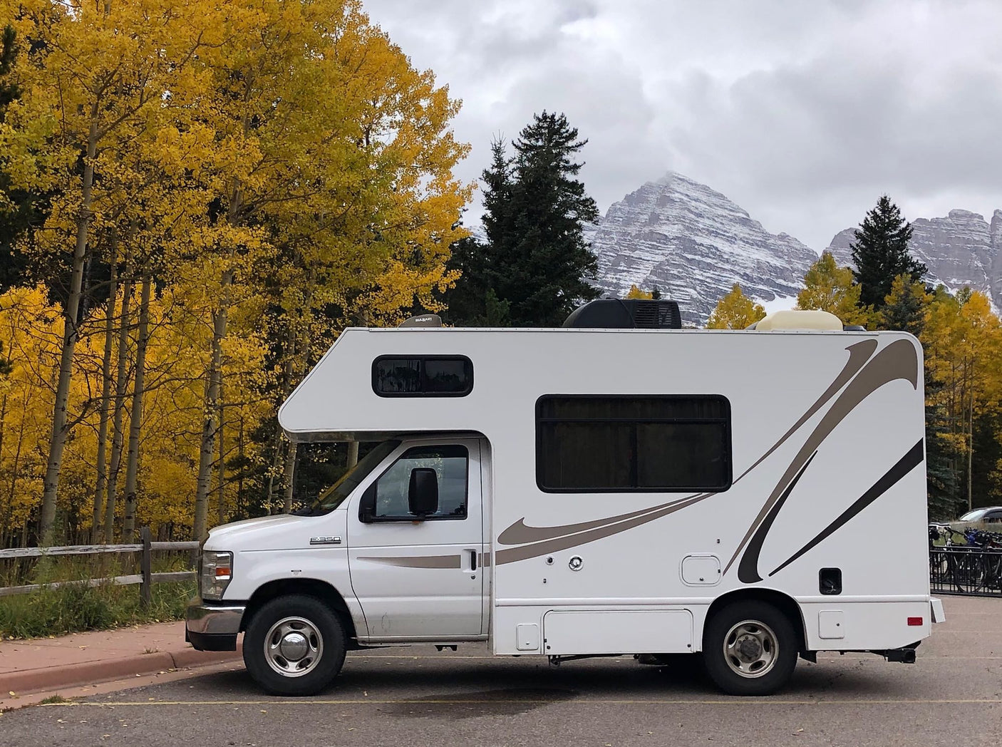 A white RV rental, a Class C motorhome, is parked among vibrant yellow autumn trees and snowy mountains under a cloudy sky.