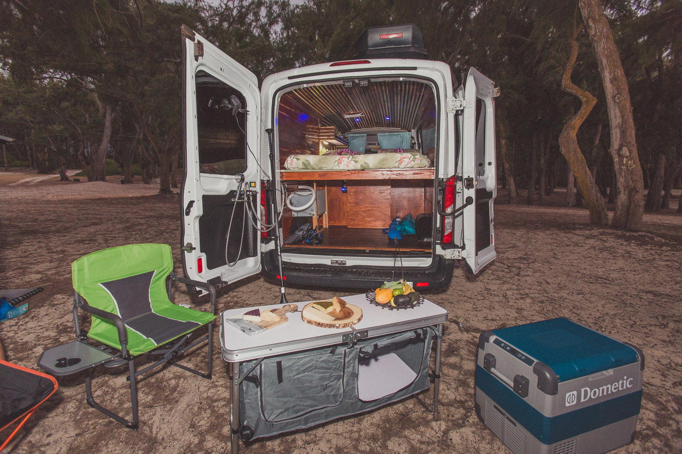 This RV rental shows an open camper van with a cozy interior, an outdoor kitchen setup with a Dometic cooler and snacks in a natural setting.