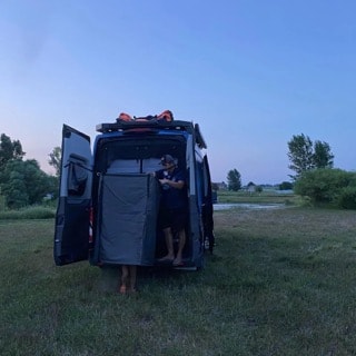 A person uses a privacy screen inside the open back of an RV rental campervan at dusk, parked in a grassy field. Ideal for camping.