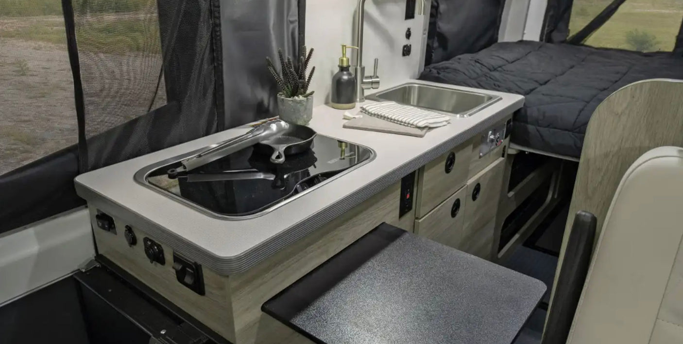 RV rental interior: Kitchenette with sink, cooktop, and pull-out table. Bed in background. Ideal for travel.